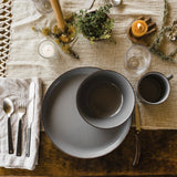 The bowl sits on top of a matching plate on a wood table set with silverware, linens, candles, herbs, glass, and a matching mug.