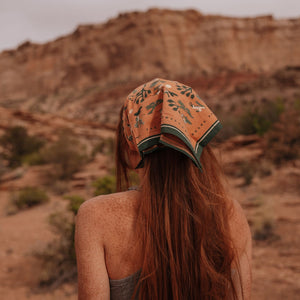 A girl with red hair wears the bandana tied around her hair. She faces away from the camera towards a desert view.