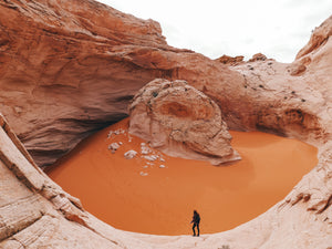 A massive natural rocky bowl fills the frame. Inside the bowl is orange desert sand and a unique dome-shaped rock formation. A girl stands a ways below the camera on the rocky ledge looking into the bowl.