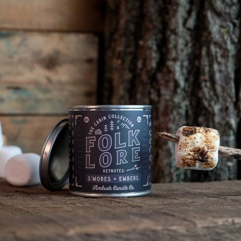 Candle labeled 'Folk Lore' with a toasted marshmallow on a stick on a wooden surface.