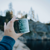 Hand holding a 'In the Pines' Candle with a forest and lake background