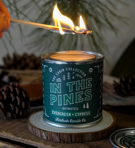 The candle labeled 'In The Pines' sitting on a wood coaster with a lit match held over the top.