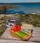 The mini olive wood board with peppers, celery, and hummus on it sitting next to a full camp mug on a picnic table in front of a lake.