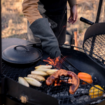 A man wearing a fire glove holds a pair of tongs and handles a pepper sitting in the coals of a fire inside a large grill. Just above the fire on a rack, there is a sausage, vegetables, and a cast iron skillet.