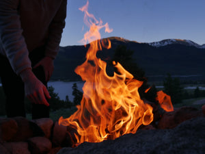 A fire burns in a rocky fire pit. A man's hand is extended towards the fire as he has just dropped wood into the flame. In the background is a pine forest, lake, and mountains with only the top of the peaks lit by the setting sun.