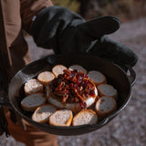 A man holds a cast iron skillet with the fire glove covering his hands. Inside the skillet is a wheel of brie covered with cranberries and walnuts surrounded by baguette rounds.