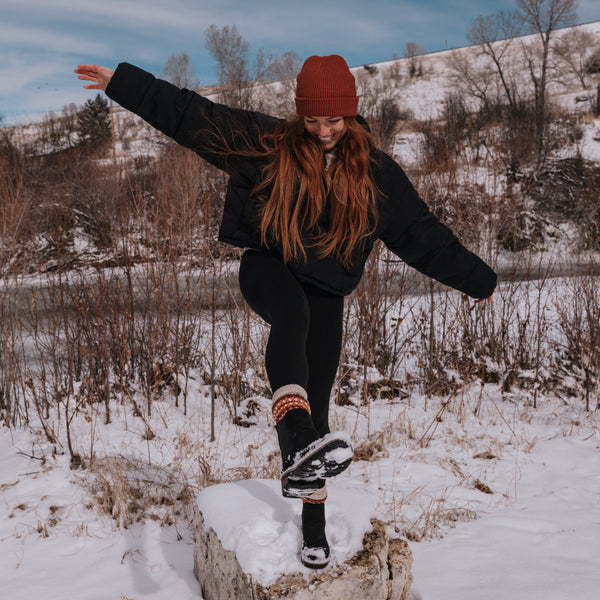 A girl with red hair wearing winter clothing, playing in the snow. Her arms are out and her leg is outstretched towards the camera, showing the Asenka Sock