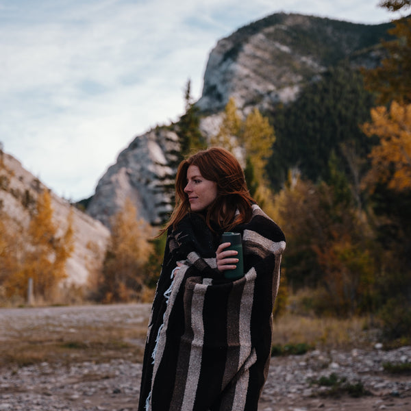A girl stands in front of a mountain view looking off to the side. She is wrapped in a blanket and holds a travel mug in her hand.