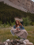 A girl  sitting in a camp chair next to a fire ring in the mountains with the Cabin Time Blanket on her lap.