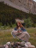 A girl  sitting in a camp chair next to a fire ring in the mountains with the Cabin Time Blanket on her lap.