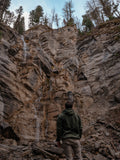 Man wearing a green jacket standing in a rocky, natural setting with a waterfall in the background