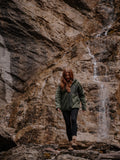 Girl wearing a green jacket standing in a rocky, natural setting with a waterfall in the background