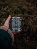 Hand holding a 'In the Pines' Candle with a green vegetation in the background