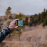Hand holding a candle tin labeled 'Scenic Route' with a field and forest in the background