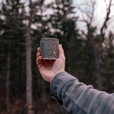 Hand holding a candle tin labeled 'Take a Hike' against a forest background