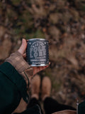 Camera view looking down a hand holding a candle tin labeled 'Folk Lore' with brown boots and grassy setting in the background 