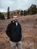 Man wearing a black jacket standing in a field with mountains and trees in the background