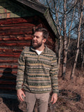 Man wearing a patterned fleece standing in front of a wooden cabin.