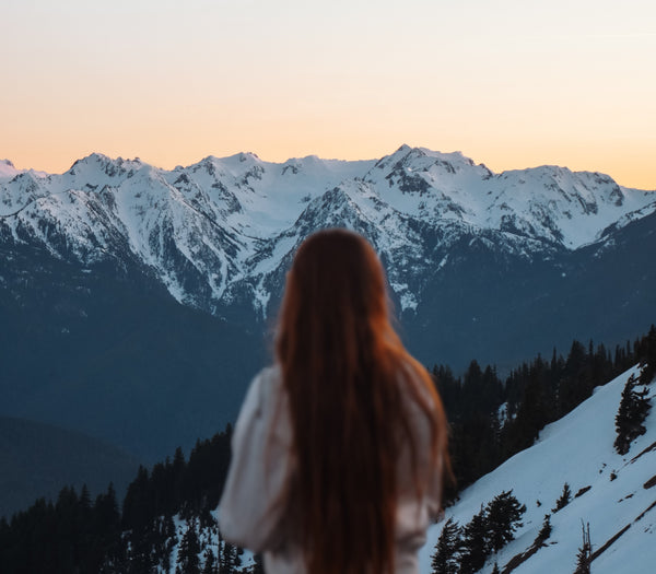 A girl stands on a mountain top facing away from the camera in front of rugged, snow-covered peaks. She is blurry, while the mountains are in focus.