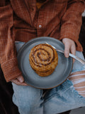 The photo looks down on a plate with a stack of pumpkin cinnamon roll pancakes and a fork on it. A girl wearing jeans and a flannel holds the plate on her lap.