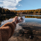 A hand is outstretched in front of the camera holding a steaming mug that reads "Please help Smokey prevent wildfires." with an image of Smokey Bear's head. Behind the mug is the view of a lake with yellow larch trees on the shore.