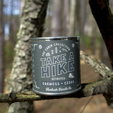 Candle canister labeled 'Take a Hike' on a branch in a forest setting