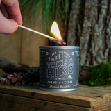 Candle labeled 'Take a Hike' being lit with a match against a natural background.