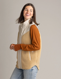 Woman standing in front of a blank white background facing to the side and smiling. She's wearing the Vela Vest over a long-sleeve and jeans.