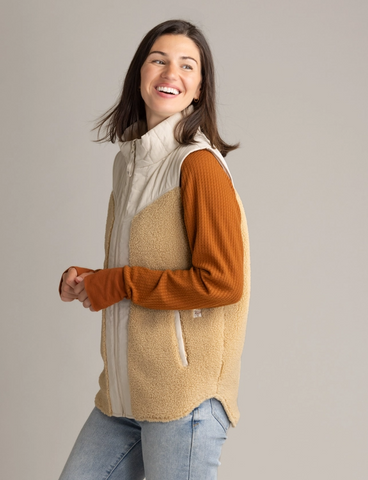 Woman standing in front of a blank white background facing to the side and smiling. She's wearing the Vela Vest over a long-sleeve and jeans.